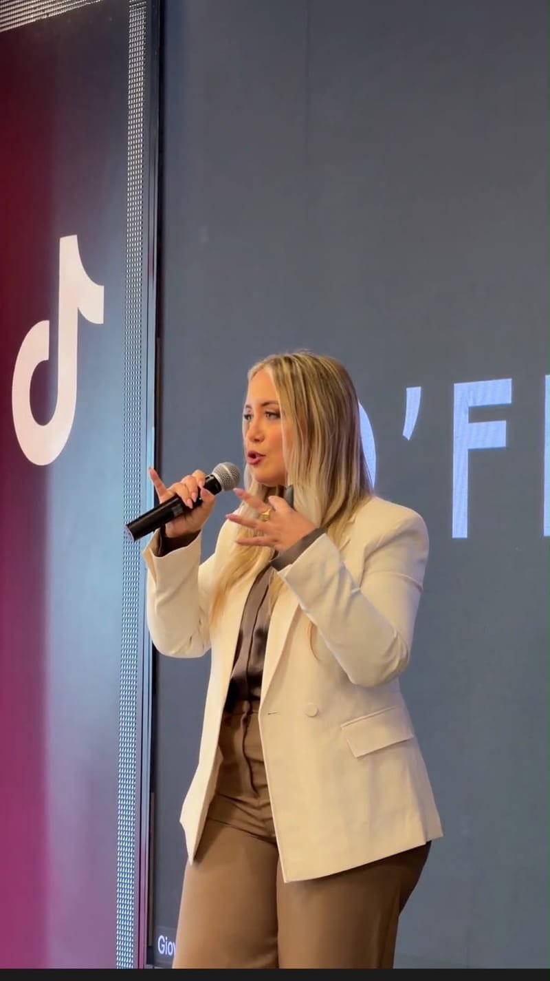 Kelly Vial falando ao microfone em um evento, com os logos do TikTok e da D’France no telão